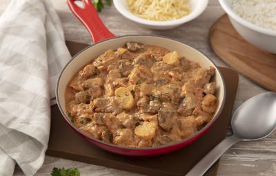 Foto da receita de strogonoff de carne. Observa-se uma panela grande com o strogonoff sobre uma tábua de madeira. Ao lado, um colher de servir e duas tigelas: uma com batata-palha e outra com arroz.