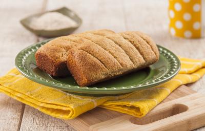 Fotografia em tons de amarelo em uma bancada de madeira clara, um pano amarelo com listras brancas, uma tábua de madeira, um prato verde redondo com dois pãezinhos em cima dele. Ao fundo, uma xícara amarela com bolinhas brancas.