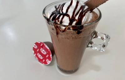 Fotografia em tons de branco com uma caneca de vidro ao centro. Dentro da caneca existe uma receita de frappuccino de cor chocolate coberto com chantilly e calda de chocolate. Ao lado existe uma cápsula de NESTLÉ Dolce Gusto KitKat