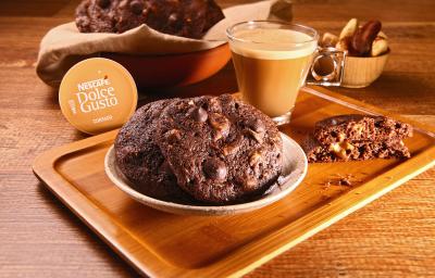 Fotografia em tons de bege em uma bancada de madeira escura, uma tábua de madeira com um pratinho bege redondo e alguns cookies de chocolate com café em cima dele. Ao lado, uma xícara de vidro com o Café Cortado Dolce Gusto e ao fundo mais cookies.