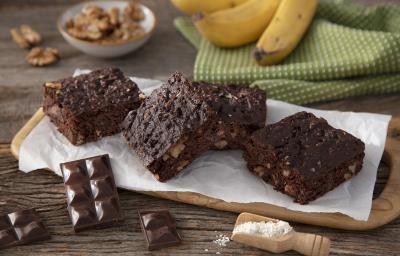 Fotografia em tons de marrom em uma bancada de madeira de cor marrom. Ao centro, uma tábua de madeira contendo os brownies em cima. Ao lado, há alguns tabletes de chocolate e ao fundo, um pano verde quadriculado com algumas bananas em cima.