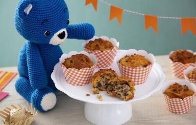 Fotografia em tons de laranja, em uma bancada de madeira de cor branca. Ao centro, uma boleira contendo os muffins com um ursinho azul, um mini chapeu de palha e um pano listrado colorido ao lado. Ao fundo, uma parede azul piscina e mini bandeiras.