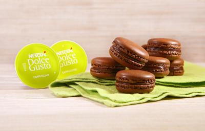 Fotografia em tons de verde em uma bancada de madeira clara, um paninho verde com vários macarons de chocolate e café em cima dele. Ao lado, cápsulas de Cappuccino Dolce Gusto.