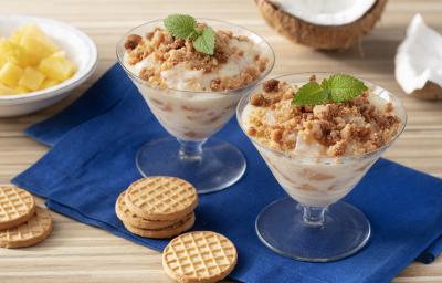 Fotografia em tons de marrom e azul de uma bancada de madeira com um paninho azul, sobre ele duas taças com o creme e biscoitos. Ao fundo um coco e um recipiente redondo com abacaxi.