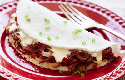 Fotografia em tons de branco e vermelho, com prato vermelho ao centro contendo uma tapioca com recheio de carne-seca, queijo de coalho e salpicada de cebolinha, acompanhada de um garfo, sobre guardanapo multicolorido.