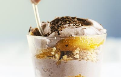 Fotografia de um copinho de vidro, dentro tem pavê com biscoitos champanhe, laranja em gomos e creme de chocolate meio amargo. Por cima do pavê tem raspas de chocolate. O copo está sobre uma toalha de mesa branca.