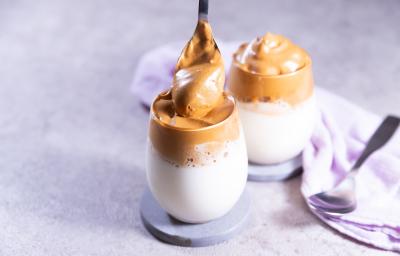 Fotografia de dois copos de vidro com cappuccino cremoso, com espuma de chocolate por cima, também cremoso. Cada copo está apoiado em um apoio cinza, o qual está sobre uma mesa de cimento queimado.