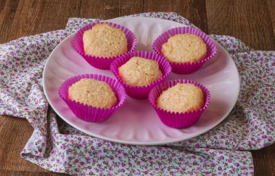 Imagem da receita de Queijadinha da Miriam Braga, servida em forminhas rosas de cupcakes, sobre um prato rosa, em um bancada de madeira decorada com um tecido florido