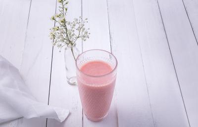 Fotografia em tons de rosa em uma bancada de madeira clara com um copo de vidro ao centro e a vitamina de hortelã com goiaba dentro dele. Ao fundo, um vasinho de vidro com flores brancas pequenas.