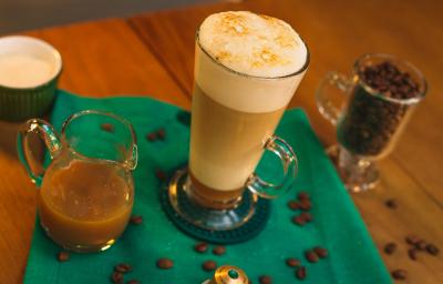 Fotografia em tons de verde de uma mesa de madeira com um paninho verde, uma xícara com a bebida e grãos de café decorando.