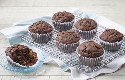 Fotografia em tons de azul em uma mesa de madeira clara com um suporte de ferro com os cupcakes integrais de cacau e banana em forminhas de papel. Ao lado, um pratinho de vidro com um cupcake cortado ao meio.