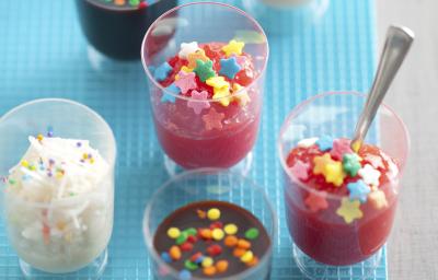 Fotografia em tons de azul em uma bancada cinza com copinhos transparentes contendo a cocadinha, o brigadeiro de colher e o docinho de morango de colher. Ao lado, confeitos coloridos espalhados em formato de estrela.