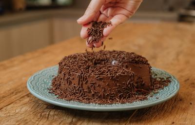 Fotografia em tons de marrom de uma bancada de madeira sobre ela um prato redondo azul com um brigadeirão. Em cima uma mão salpicando granulado de chocolate.