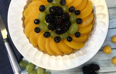 Foto vista de cima da receita de Torta de Frutas com Leite Moça, decorada com pêssegos, uvas, blueberrie, sobre um prato branco em uma bancada de madeira.