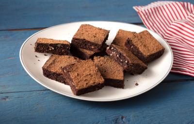 Fotografia em tons de azul, branco e  marrom de uma bancada azul com um prato redondo branco, sobre ele pedaços de brownie de batata-doce. Ao lado um paninho listrado vermelho e branco.