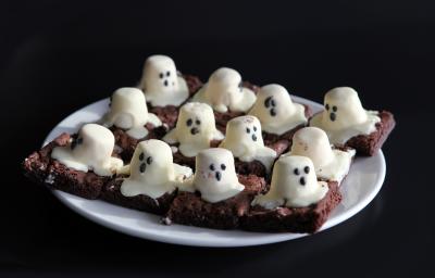 Fotografia de um prato branco com pedaços de brownie de chocolate cortados em quadrados, com marshmallows derretidos em cima representando um fantasma.