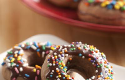 Fotografia em tons de marrom, branco e vermelho de uma bancada de madeira, sobre ela um prato branco com um donuts. Ao fundo um prato vermelho com donuts.