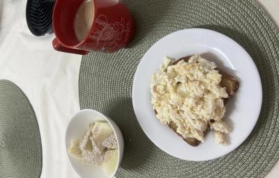 Fotografia de um prato raso médio branco com uma fatia de pão e ovos mexidos cremosos com farelo de aveia. Ao lado tem um recipiente pequeno de vidro e branco com melão e aveia e uma xícara vermelha grande.