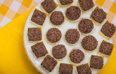 Fotografia em tons de amarelo em uma bancada de madeira com um pano amarelo e outro amarelo xadrez com branco. Em cima deles, um suporte para bolo branco com vários brigadeiros em forminhas amarelas.