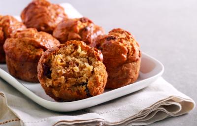 Fotografia de alguns muffins de maçã sobre um apoio retangular branco, o qual está apoiado em um paninho também branco. O pano está sobre uma bancada de cimento queimado claro.