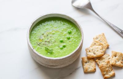 Fotografia em tons de verde em uma bancada de madeira de cor branca. Ao centro, uma tigela contendo a sopa de ervilha com alguns biscoitos de água e sal e uma colher ao lado.