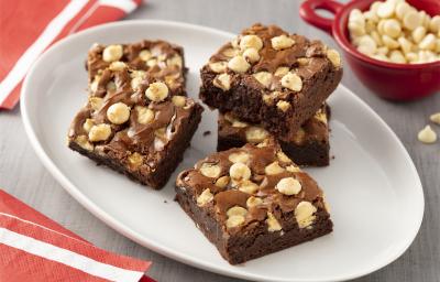 Fotografia em tons de branco, vermelho e cinza de de uma bancada cinza com um prato oval branco, sobre ele cinco pedaços de brownie. Ao fundo um recipiente redondo com gotas de chocolate branco.