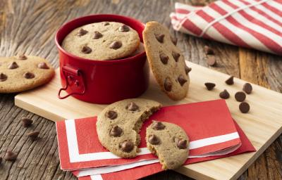 Fotografia em tons de vermelho em uma bancada de madeira com uma tábua de madeira, um guardanapo de papel vermelho com listra branca, um cookie com gotas de chocolate cortado ao meio em cima dele. Ao fundo, uma panelinha vermelha pequena com mais cookies.