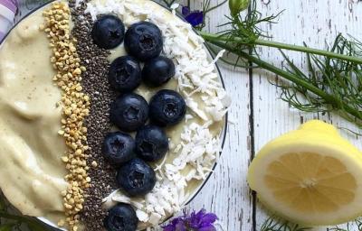 Fotografia em tons de roxo em uma bancada de madeira com um bowl e o creme de avocado dentro dele, decorado com mirtilo, castanha-de-caju e sementes de chia. Ao lado, limão siciliano na mesa.