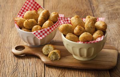 Fotografia em tons de marrom, com dois bowls de porcelana forrados com guardanapos quadriculados em vermelho e branco, sendo um sobre tábua com um bolinho aberto, ambos com bolinhos de macaxeira fritos inteiros, tudo sobre bancada de madeira rústica.