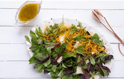 Fotografia em tons de branco e verde, ao centro recipiente com folhas verdes, roxas e tiras de abóbora, ao lado pegador de salada e potinho com molho, tudo sobre bancada na cor branca.