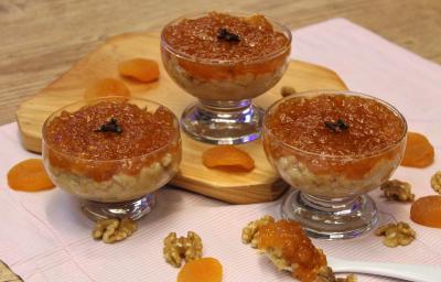 Fotografia em tons de laranja em uma bancada de madeira de cor marrom. Ao centro, uma toalha rosa com uma tábua de madeira. Em cima, estão as taças de brigadeiro e ao redor, damascos.