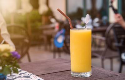 Fotografia de um copo grande com um refresco de laranja, dentro do copo, um canudo de cor marrom e um enfeito de vidro. O copo está sobre uma mesa de madeira na cor marrom. Ao fundo, mesas e cadeiras de uma cafeteria ao ar livre.