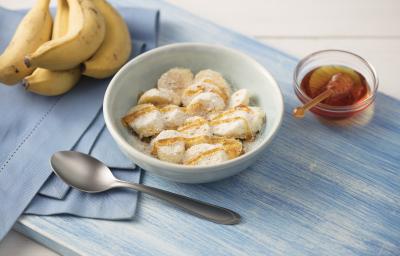 Fotografia em tons de azul em uma mesa de madeira clara com um paninho azul ao lado, um prato branco redondo fundo com as rodelas de banana cobertas com mel, aveia, leite em pó e linhaça. Ao lado, um cacho de bananas e um potinho com mel.