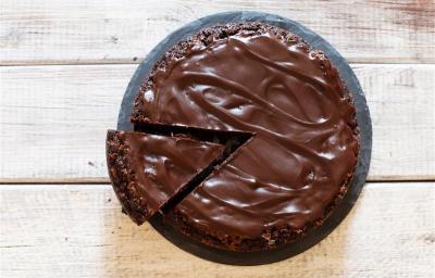 Fotografia em tons de marrom e branco de uma bancada marrom vista de cima, contém um prato redondo e branco com um bolo de chocolate com cobertura de chocolate e uma fatia sendo retirada por uma espátula