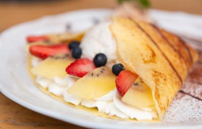 Fotografia em tons de branco com um prato branco ao centro. Em cima do prato existe um crepe recheado com um creme branco doce, morango, kiwi e blueberries.