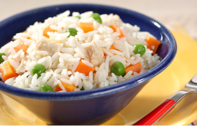 fotografia em tons de amarelo, azul e branco tirada de um prato redondo amarelo com um recipiente azul, que contém o arroz com pedaços de cenouras e ervilhas com um garfo ao lado para servir