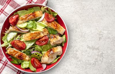 Fotografia vista de cima de um salada de folhas pepino, tomate, frango, cebola roxa e salsinha com um molho de maracujá e mel. A salada está dentro de um recipiente fundo vermelho, apoiado sobre e metade de um pano branco com linhas vermelhas.