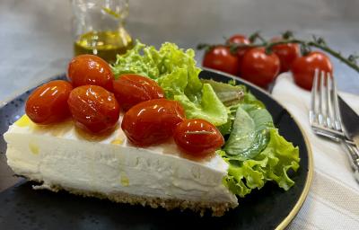 Foto da receita de Cheesecake Salgado. Observa-se uma fatia da torta com tomatinhos confit em cima em um prato preto servido com uma salada de alface.