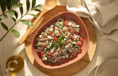 Foto da receita de tabule de aveia corte rústico servida em um prato fundo rosa salmão sobre uma tábua de madeira circular com um garfo de madeira ao lado e um pano de linho