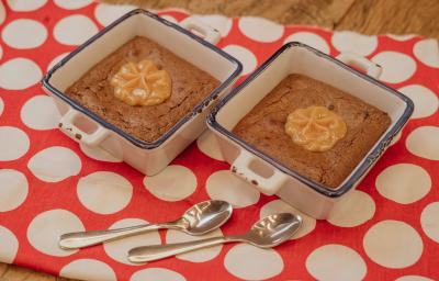 Fotografia em tons de marrom e vermelho de uma bancada de madeira sobre ela um paninho vermelho com bolinhas brancas com duas colheres se sobremesa. Sobre ele dois recipientes quadrados e brancos, dentro o brownie e o MOÇA doce de leite.