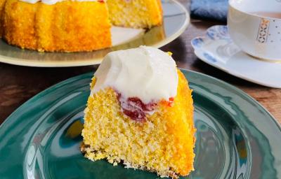 Fotografia em tons de verde e amarelo com um prato verde ao centro. Em cima do prato existe uma fatia de bolo de fubá com cobertura de goiabada e cream cheese. Ao fundo existe o bolo inteiro com uma fatia cortada