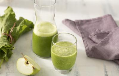 Fotografia em tons de verde e roxo de uma bancada branca, sobre ela dois copos de vidro com suco verde. Ao lado um paninho roxo, uma fatia de maçã verde e folhas de couve.