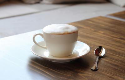 fotografia em tons de marrom e branco tirada de uma bancada marrom. Contém um pratinho branco com uma xicara de café branca, e dentro contém a bebida Cappuccino.