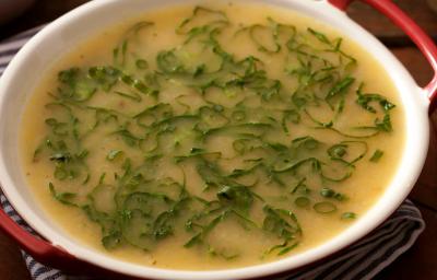 Fotografia em tons de marrom e branco de uma bancada de madeira com paninho listrado azul e branco sobre ele uma panela com sopa de mandioca com couve.