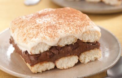 Fotografia em tons de bege em uma mesa de madeira com uma toalha amarela e um prato bege redondo raso ao centro com um pedaço de pavê de chocolate.