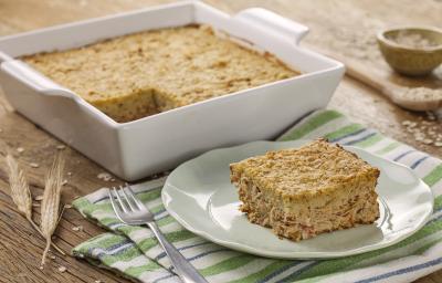 Fotografia em tons de branco e dourado, refratário branco com torta de frango, prato branco com uma porção de torta de frango com um garfo ao lado e sobre guardanapo com listras verdes, azuis e brancas, colher e potinho com aveia ao fundo.