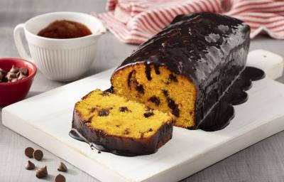 Fotografia em tons de branco e vermelho de uma bancada branca branca com um prato retangular branco, sobre ele um bolo de cenoura. Ao fundo um paninho listrado vermelho e branco e um recipiente redondo branco com calda de chocolate.