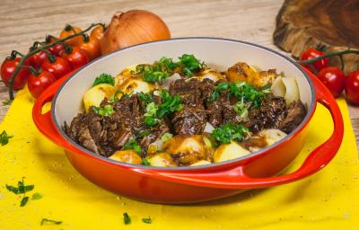 Foto aproximada de uma travessa vermelha com a receita de Carne de Panela, em uma bancada de madeira decorada com cebola, tomate-cereja, um tecido amarelo e algumas cebolinhas verdes