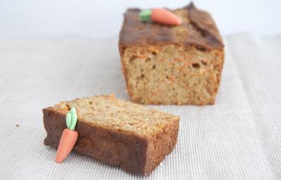 Fotografia em tons claros de um bolo de cenoura com mini cenouras em cima, feitas de pasta americana, cortado em uma fatia grossa que está mais no centro da foto. O bolo está sobre uma toalha de mesa na cor marrom claro.