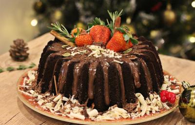 Fotografia em tons de marrom e vermelho de uma bancada de madeira clara, sobre ela um prato vermelho redondo com o bolo de chocolate decorado. Ao lado uma bolinha natalina e uma pinha.